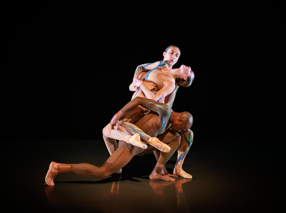a woman in sheer leotard and tights, seams tangled in the center of two men.